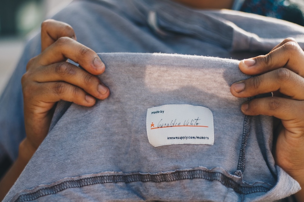 A Known Supply garment maker holds up a piece of clothing they made with their signature tag stitched in.
