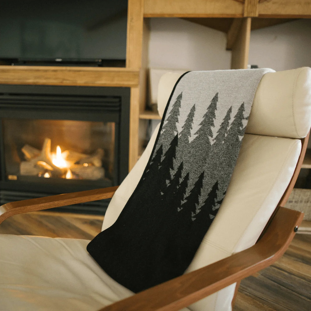 A Tentree Organic Cotton Juniper Blanket draped over a white chair in front of a cozy fireplace, can be designed and curated by Swagger.