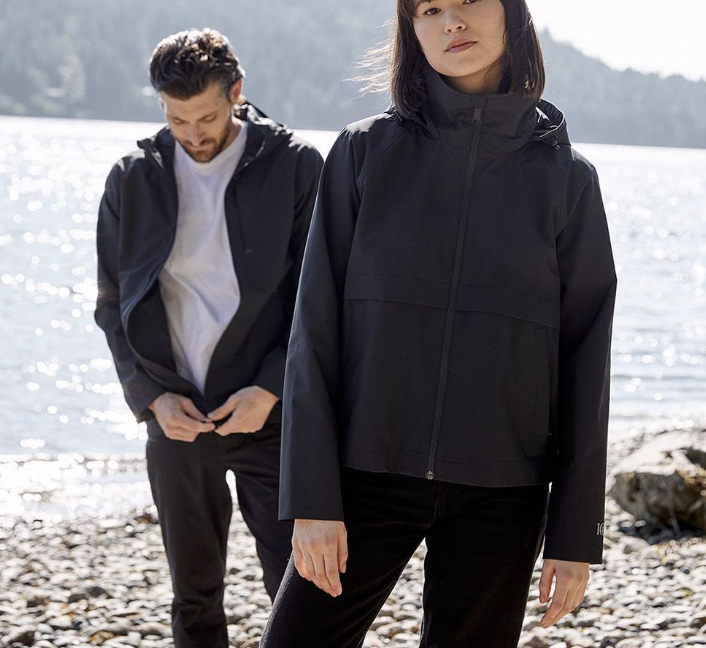 Two people standing on the shore wearing the customized Tentree Nimbus Long Waterproof Rain Jacket by Swagger.