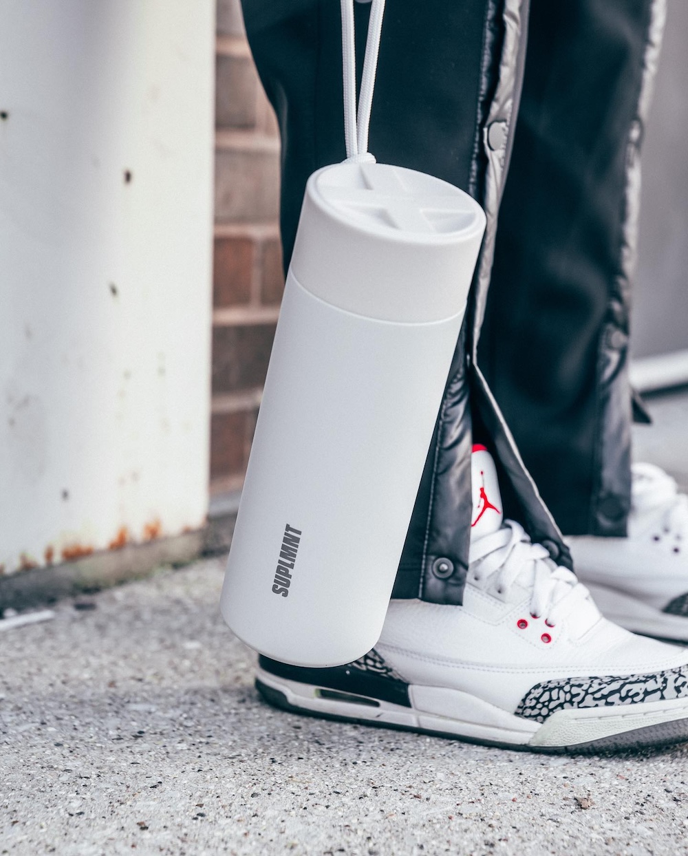 Image of person holding SUPLMNT 24 Oz Insulated Water Bottle With Classic Lid in white in front of leather pants and Air Jordan shoes.
