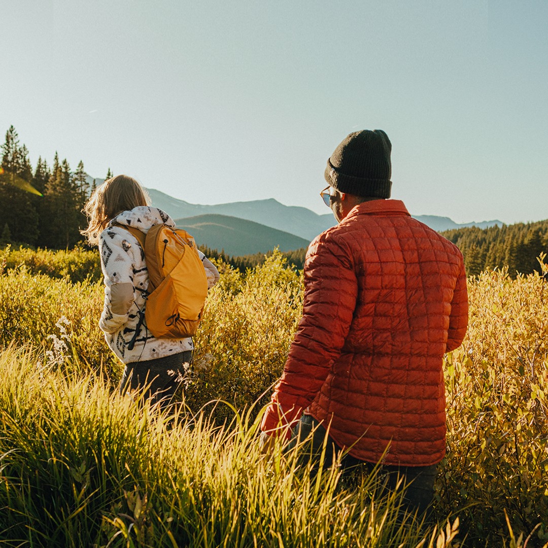 Woman with The North Face yellow backpack and man with orange The North Face Thermoball Trekker Jacket walking through a grass field with mountains in the backround.