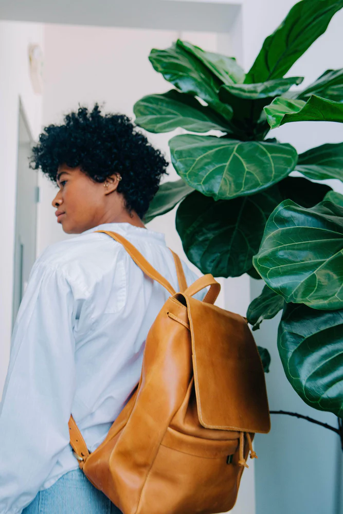 Woman stands by a fig tree while wearing the Enku Leather Backpack can be custom branded by Swagger.
